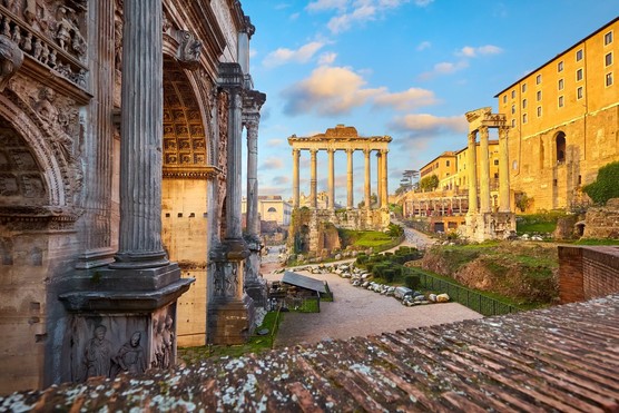 An image of Roman ruins, including tall structures with 6 columns in the background. There is a large curved wall in the foreground