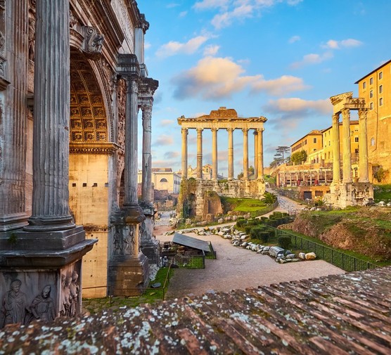 An image of Roman ruins, including tall structures with 6 columns in the background. There is a large curved wall in the foreground