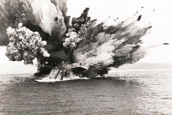 A black and white photograph of a large, smoky explosion on the sea