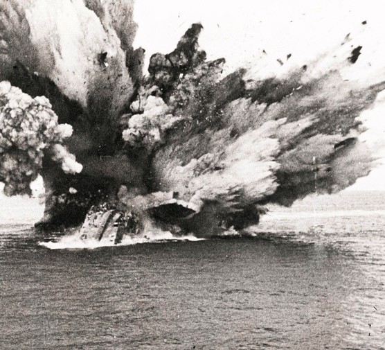 A black and white photograph of a large, smoky explosion on the sea