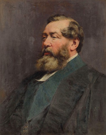 A man sits facing the left. He has a thick beard and brown hair and has a blue collared jacket