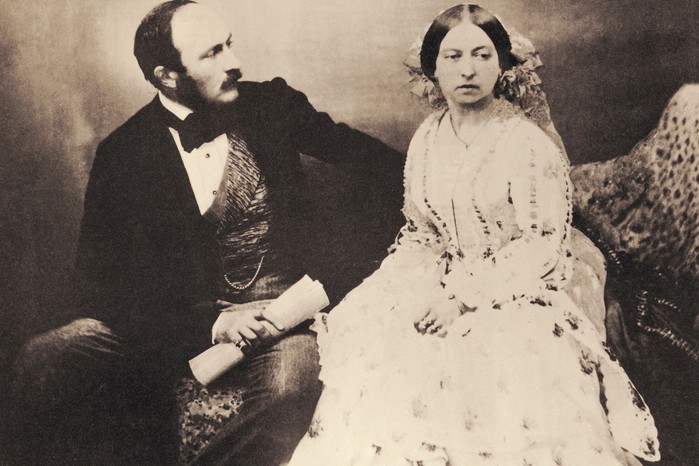 A black and white photograph of Victoria and Albert. He sits on the left, dressed in a black suit and bow tie, with one arm leaning on the back of the seat and the other holding a scroll. Victoria, dressed in a long white dress, sits on the right, looking slightly away from Albert and the camera