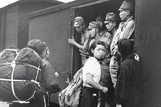 A black and white photograph shows a group of Japanese military men on a train, while some young women prepare to get on the carriage. A young woman looks back over her shoulder