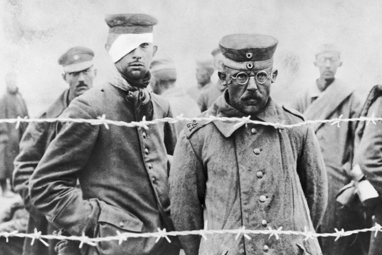 Two men dressed in military uniform stand behind a barbed wire, looking into the camera. One wears a bandage around his head, covering one eye