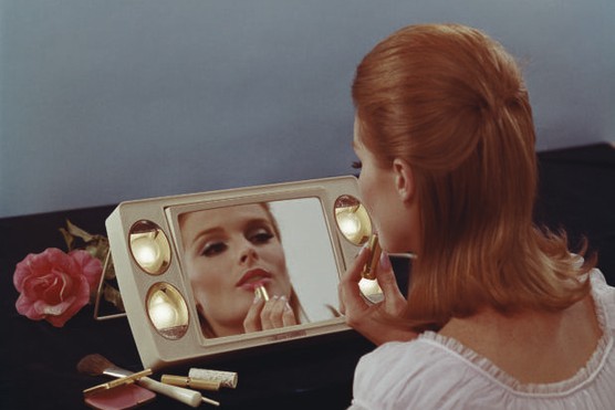 A young woman sat in front of a mirror applies lipstick, 1967