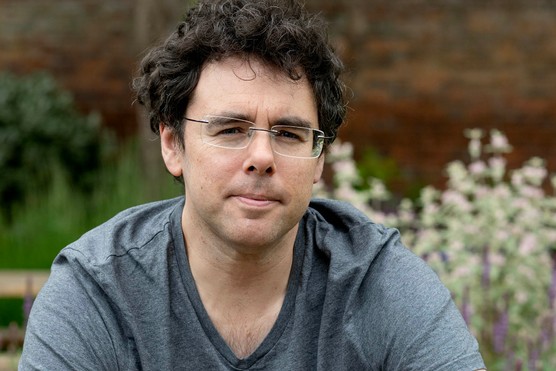 A man in a grey T shirt and blue jeans sits with his arms crossed, facing the camera. He has dark curly hair and wears glasses
