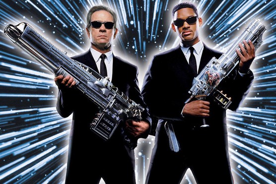 Two men stand next to each other holding large guns. They are dressed in black suits and ties with sunglasses and are standing against a blue and black background. Above them, the words "MIB: MEN IN BLACK" are written