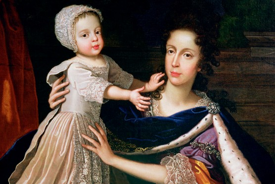 A woman dressed in a ruffled dress and spotted robe holds a young child in a white, lacy dress and cap