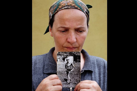 A woman dressed in a grey top and a green and orange bandana holds a black and white photograph of a young man against her chin. She stands in front of a plain, yellow wall