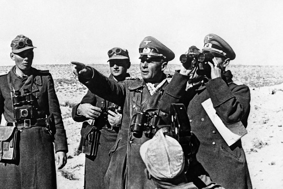 A black and white photograph of three men in military uniform. The man in the middle, is holding his arm out pointing to something in the distance and to his left, is a man using binoculars