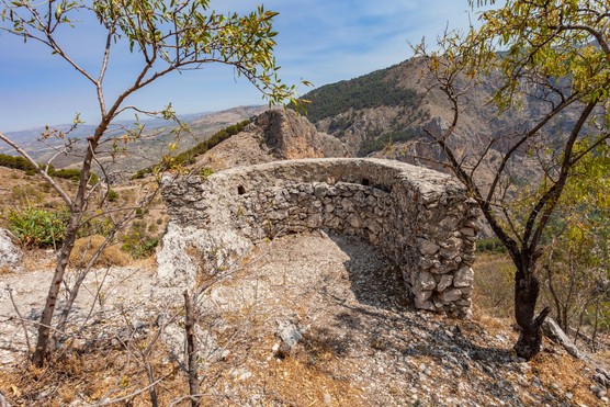 A landscape image shows a grey and orange stone wall in a semi-circle shape, overlooking a large hill range and a blue sky. On either side of it there are trees