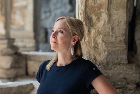 A woman with blonde hair and wearing a navy T shirt stands looking up towards the top left of the image. She has a large white earring and stands in front of some Roman ruins